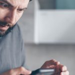 Homem barbado demonstra surpresa enquanto olha para o smartphone em um ambiente interno.