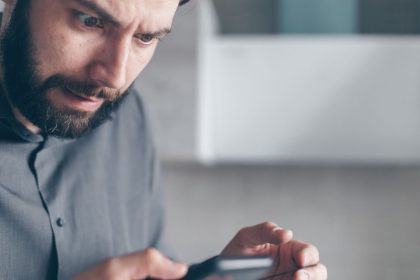 Homem barbado demonstra surpresa enquanto olha para o smartphone em um ambiente interno.