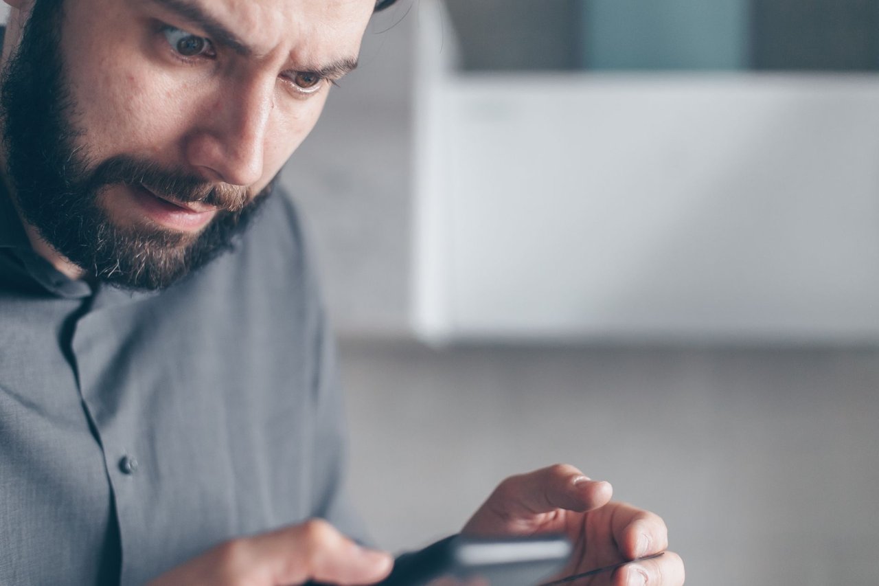 Homem barbado demonstra surpresa enquanto olha para o smartphone em um ambiente interno.